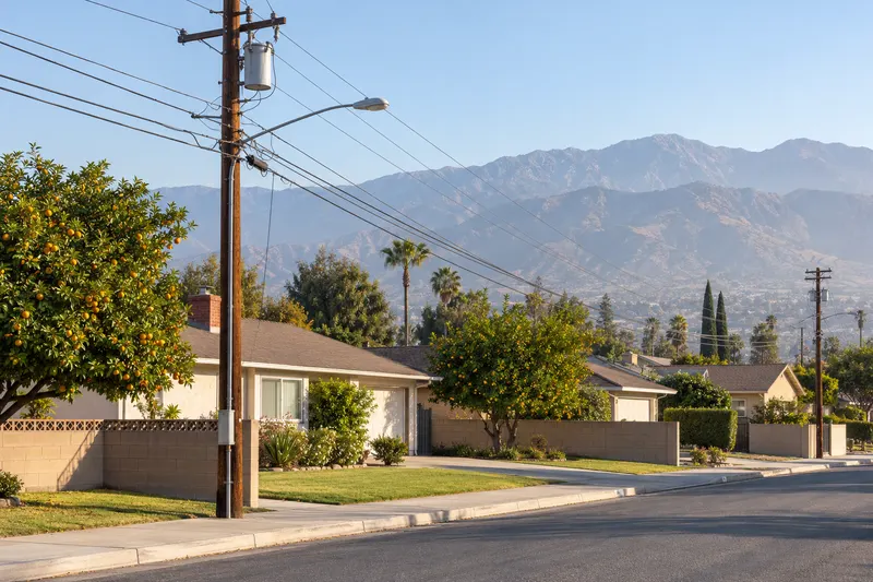 Highland, California: a street of 1970s single-story stucco homes with citrus trees and a utility pole, mountains behind